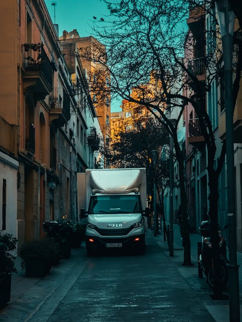 A white Iveco moving van is positioned on a narrow urban street, with buildings on either side and leafless trees overhead. The van is in the process of a home relocation, with the rear doors closed, indicating it is either preparing to leave or has just arrived. The street is lined with black bollards, and there are a few visible packages or boxes on the pavement near the van, suggesting packing and furniture transport activity. The scene is illuminated by late afternoon or early evening natural light, casting shadows across the street, which is typical during a domestic moving or packing and moving process. The presence of the van and the urban environment underlines the logistical aspect of commercial and residential removal services, as provided by Harrow Man and Van, supporting efficient loading and transportation operations for house removals within the city.