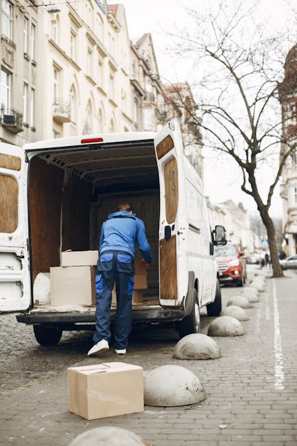A white Iveco moving van is positioned on a narrow urban street, with buildings on either side and leafless trees overhead. The van is in the process of a home relocation, with the rear doors closed, indicating it is either preparing to leave or has just arrived. The street is lined with black bollards, and there are a few visible packages or boxes on the pavement near the van, suggesting packing and furniture transport activity. The scene is illuminated by late afternoon or early evening natural light, casting shadows across the street, which is typical during a domestic moving or packing and moving process. The presence of the van and the urban environment underlines the logistical aspect of commercial and residential removal services, as provided by Harrow Man and Van, supporting efficient loading and transportation operations for house removals within the city.