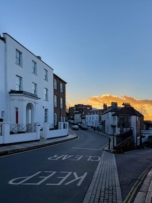 A quiet residential street during sunset with a clear blue sky and orange clouds, featuring a row of white and dark-colored terraced houses with traditional architectural details such as bay windows and decorative trim. The street is gently curved with a marked 'KEEP CLEAR' sign painted on the asphalt near the curb, which is made of grey paving slabs. A sidewalk runs alongside the road, separated by a curb, and is lined with bollards and tactile paving for pedestrians. The scene is well-lit by natural evening light, highlighting the cleanliness and order of the area. This image depicts a typical UK urban street scene suitable for house removals or home relocation services, exemplifying the environment in which professional removal companies like Harrow Man and Van operate when transporting furniture and belongings, often involving packing, loading, and navigating residential roads during planning and execution of moves. The setting emphasizes the importance of obtaining proper permits for parking and loading when carrying out house removals in such neighborhoods.