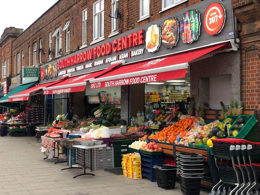 Photograph of a street-facing view of South Harrow Food Centre, a local grocery store, with a red and white awning displaying the shop's name and various food categories. In front of the store, an outdoor fruit and vegetable market is set up with several displays of fresh produce including apples, oranges, watermelons, and other fruits, along with vegetables such as cabbages and onions, arranged in baskets and crates. There are additional items like plastic crates, shopping trolleys, and cardboard boxes stacked nearby. To the left, a green sign indicates a fresh fruit and vegetable section. The building is constructed with brick, and the scene is illuminated by daylight, suggesting daytime activity. This setting depicts typical logistics involved in home relocations or moving processes, where packing, loading, and transport preparations may take place in a commercial environment such as this, relevant to removals and transportation services offered by Harrow Man and Van.