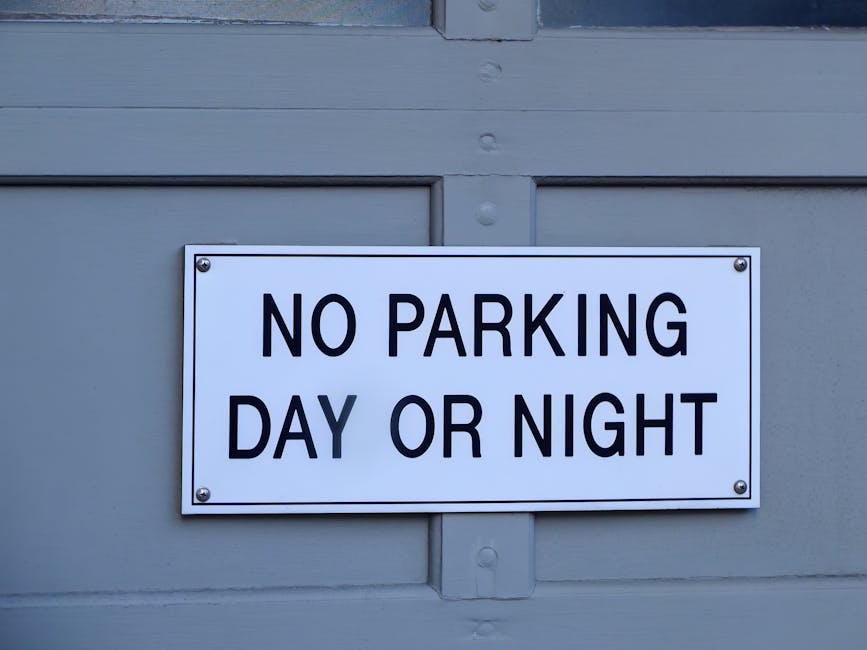 A close-up photograph of a rectangular white sign with bold black text reading 'NO PARKING DAY OR NIGHT' mounted on a grey wooden surface, likely a garage door or exterior wall. The sign is attached with four metallic screws, one at each corner, and is positioned in the middle of the image. Behind the sign, the grey surface shows horizontal paneling with visible wood grain and some textured details. The lighting is neutral, illuminating the sign clearly for visibility. This image relates to housing and property management, indicating parking restrictions in residential or commercial areas, which may be relevant for house removals and moving services provided by Harrow Man and Van, especially when considering parking regulations on moving day.