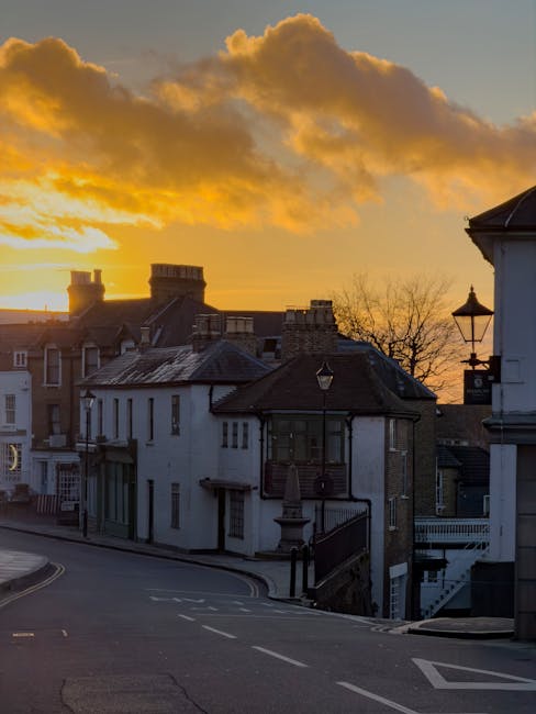 Photograph showing a quiet street in Harrow on the Hill during sunset, with the sky illuminated by orange and yellow clouds. The scene includes residential buildings with various architectural styles, including brick and white-painted facades, some with chimneys. The street is empty, paved, and features a slight curve, with a black wrought-iron fence and street lamps along the sidewalk. In the background, the silhouette of rooftops and chimney stacks is visible against the vibrant sky. This image, related to house removals and moving services, highlights the characteristic narrow streets often navigated during home relocations in Harrow on the Hill. Such surroundings necessitate careful planning and the use of appropriate furniture transport and loading processes, which companies like Harrow Man and Van manage professionally.