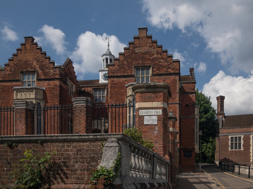 Photograph of a historic red brick building situated on Church Hill, with a decorative stepped gable roofline, tall narrow windows, and a white clock tower with a weather vane on top, set against a partly cloudy blue sky. In the foreground, a stone and brick boundary wall with a black wrought iron fence separates the street from the property. The wall features a white street sign reading 'Church Hill' mounted on a brick column. A small green shrub is visible at the base of the fence. The scene captures a residential or institutional building, typical of a traditional UK house or church, with surrounding pavements and narrow streets indicative of a tight urban setting. The image is relevant for topics related to house removals, moving tips, or furniture transport in historic or urban environments, as provided by Harrow Man and Van.