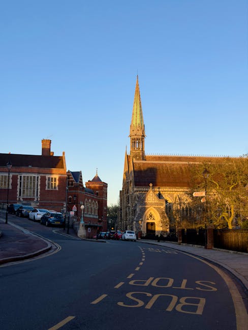 Photograph showing a quiet street in Harrow on the Hill during sunset, with the sky illuminated by orange and yellow clouds. The scene includes residential buildings with various architectural styles, including brick and white-painted facades, some with chimneys. The street is empty, paved, and features a slight curve, with a black wrought-iron fence and street lamps along the sidewalk. In the background, the silhouette of rooftops and chimney stacks is visible against the vibrant sky. This image, related to house removals and moving services, highlights the characteristic narrow streets often navigated during home relocations in Harrow on the Hill. Such surroundings necessitate careful planning and the use of appropriate furniture transport and loading processes, which companies like Harrow Man and Van manage professionally.