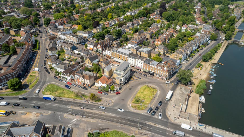 An aerial photograph of North Harrow showing a densely built residential area with rows of terraced and semi-detached houses, interspersed with trees and green spaces. The image features a river or canal on the right side, with a small dock area and boats moored along the water’s edge. In the foreground, there is a busy main road with multiple vehicles, including cars, vans, and a blue delivery truck, some moving and others parked along the street. A roundabout is visible, connecting various roads leading into the neighbourhood. Near the waterfront, there is an open paved area with several trucks and vans parked, signifying a loading or unloading zone associated with home relocation or furniture transport activities. The overall scene highlights the typical urban landscape of North Harrow, with clear sunlight illuminating the scene and shadows cast by trees and buildings, emphasizing the movement of household goods and furniture on local streets, which aligns with the services provided by Harrow Man and Van in house removals and moving logistics.