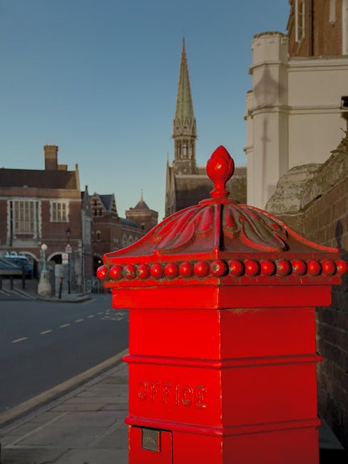 A bright red post box with decorative bead detailing on its rounded, domed lid is situated on a sidewalk near a brick wall. The box is marked with the word 'OFFICE' embossed on its front. In the background, there is a street scene with parked cars, historic brick buildings, and a tall, pointed church spire against a clear blue sky. The image is taken during daylight, with natural light illuminating the scene, showcasing the urban setting typical of house removals and furniture transport processes. The surroundings suggest a residential or central area suitable for home relocation efforts, which Harrow Man and Van services cover in North Harrow, including same-day removals and relocation logistics.