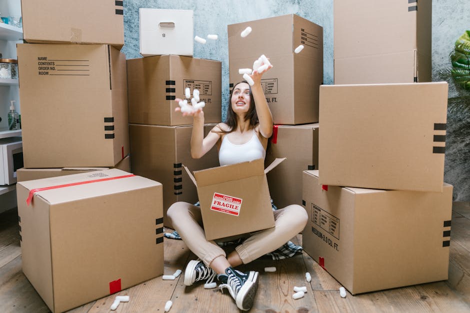 A young woman with long dark hair, sitting cross-legged on a wooden floor inside a home, surrounded by numerous unpacked cardboard boxes of various sizes, some sealed with red tape and others open revealing packing materials. She is wearing a white sleeveless top, beige pants, and sneakers, and appears to be in the process of packing or unpacking. She is smiling and tossing white foam packing peanuts into the air, with some falling into an open box placed on her lap. The background features a textured wall in a home environment, with a plant visible on the right side, and a glimpse of a kitchen counter with a few items on it. This scene depicts the packing and unpacking stage of a home relocation, consistent with professional removals services like those offered by Harrow Man and Van, focusing on furniture transport and moving logistics.