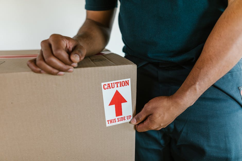 A young woman with long dark hair, sitting cross-legged on a wooden floor inside a home, surrounded by numerous unpacked cardboard boxes of various sizes, some sealed with red tape and others open revealing packing materials. She is wearing a white sleeveless top, beige pants, and sneakers, and appears to be in the process of packing or unpacking. She is smiling and tossing white foam packing peanuts into the air, with some falling into an open box placed on her lap. The background features a textured wall in a home environment, with a plant visible on the right side, and a glimpse of a kitchen counter with a few items on it. This scene depicts the packing and unpacking stage of a home relocation, consistent with professional removals services like those offered by Harrow Man and Van, focusing on furniture transport and moving logistics.