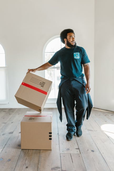 A man with dark curly hair and a beard is standing inside a bright, minimally furnished room with white walls and large arched windows, holding a cardboard box in his right hand. He is dressed in a navy blue T-shirt and dark trousers, with a jacket tied around his waist. The room features light wood flooring and appears to be part of a home undergoing packing and moving, as indicated by the presence of two sealed cardboard boxes on the floor, one of which is being lifted for furniture transport or home relocation. The natural daylight coming through the windows illuminates the space, and the scene depicts the packing and loading process typical of house removals, possibly carried out by a local moving service such as Harrow Man and Van. The overall setting suggests an organized and professional approach to packing and moving for flats or houses, consistent with the services highlighted on the page about Wealdstone man and van packing and moving for flats.