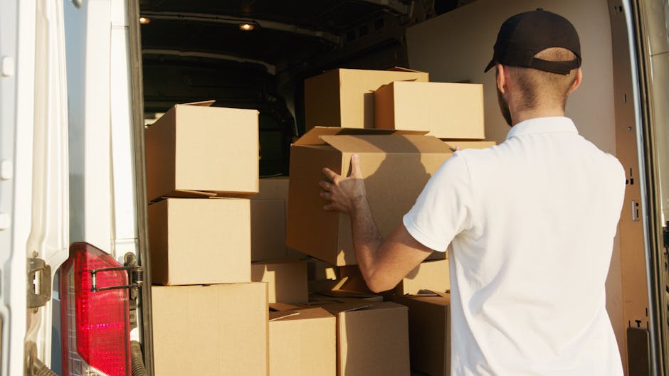 A man wearing a white shirt and a black cap is standing inside the open back of a white moving van, which is parked on a pavement. He is holding a large cardboard box, preparing to load it onto the vehicle. The van is filled with several other cardboard boxes of various sizes, some stacked on top of each other, and surrounded by packaging materials such as brown paper and bubble wrap. Additional equipment, like straps or blankets, may be visible inside the van to secure items during a home relocation. The scene captures the process of packing and moving furniture and boxes, typical of professional household removals, with natural daylight illuminating the interior and exterior environment. This image is relevant to house removal services, such as those offered by Harrow Man and Van, specialising in packing and transport for flats and houses in the Wealdstone area.